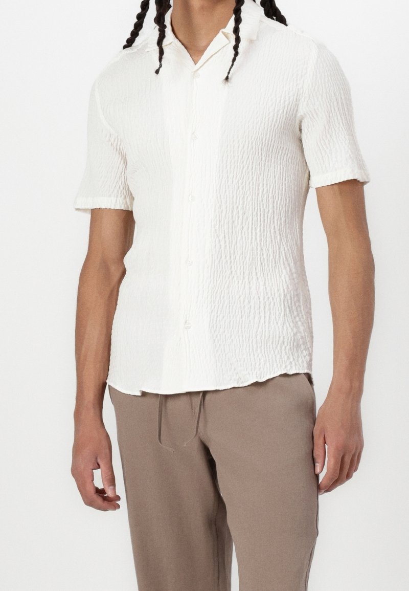 Biała koszula z krótkim rękawem z teksturowanej tkaniny, z przodu zapinana na guziki i z kołnierzykiem wykończonym w szpic, zestawiona z beżowymi spodniami.