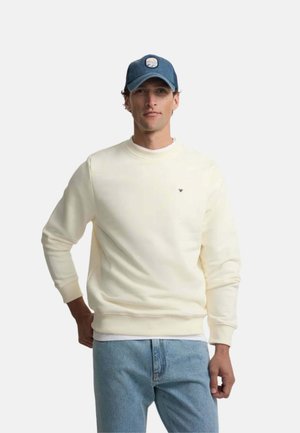 Hombre con sudadera crema de manga larga, vaqueros azules y gorra de béisbol azul marino, de pie frente a un fondo liso.