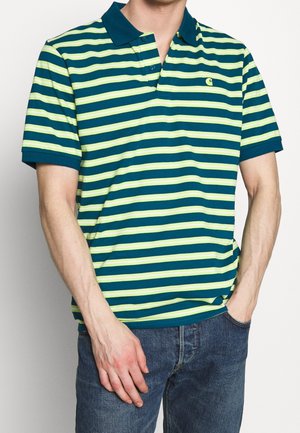 Mann trägt ein grün-gelb gestreiftes kurzärmeliges Poloshirt mit blauem Kragen und blaue Jeans, eine Hand in der Tasche.