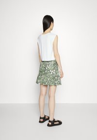 Top branco sem mangas combinado com uma saia rodada verde com padrão floral, sandálias planas pretas, visto de costas. Textura de tecido suave.