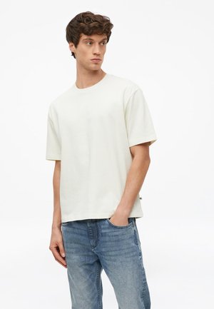 Junger Mann trägt ein schlichtes weißes T-Shirt mit kurzen Ärmeln und blaue Jeans, steht mit einer Hand in der Tasche und schaut zur Seite.