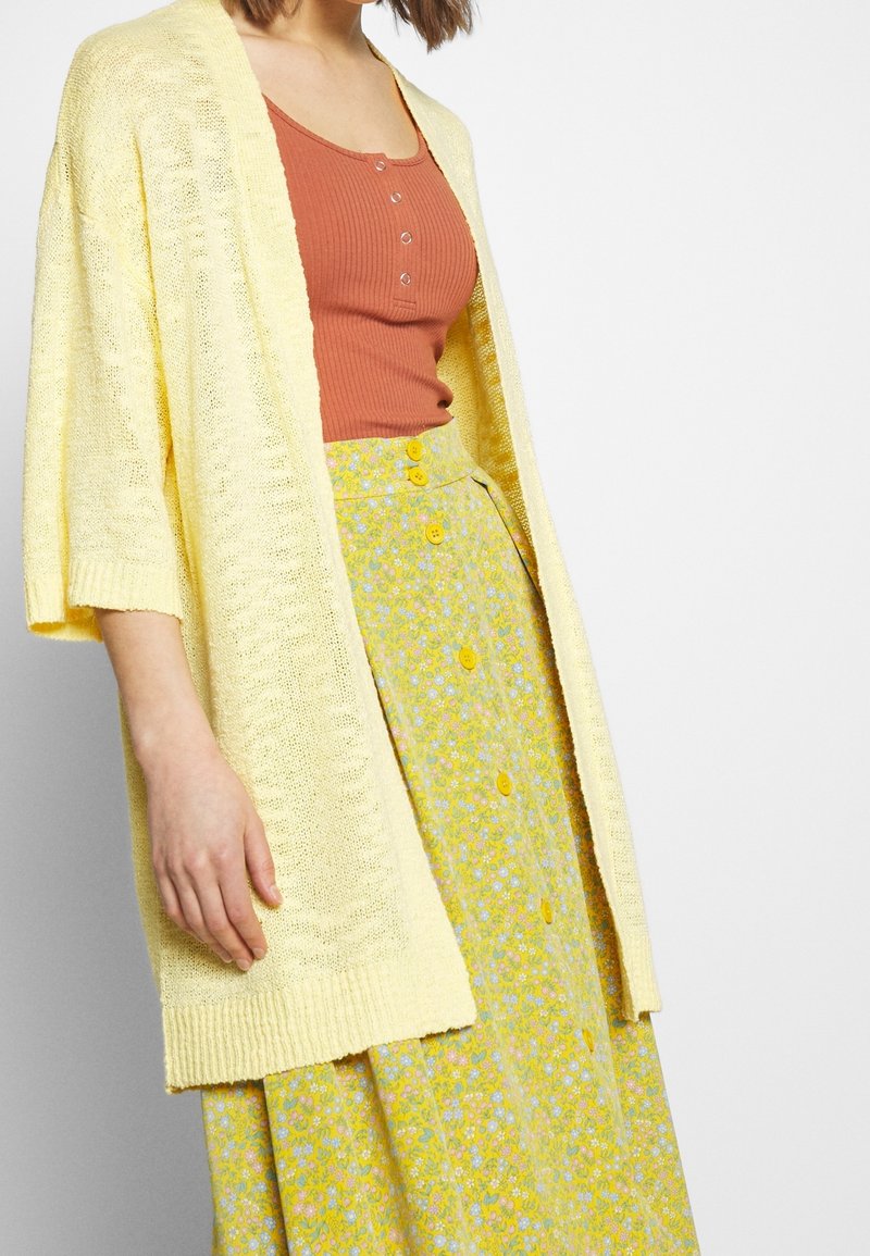 Cardigan jaune en tricot à manches larges porté sur un haut côtelé couleur rouille et une jupe florale jaune avec des accents de boutons.