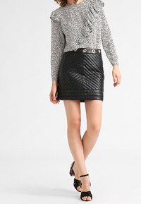 Mini gonna nera in pelle trapuntata con vita alta e bottoni decorativi. La modella indossa una blusa bianca a pois e tacchi neri.