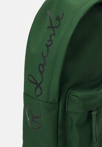 Sac à dos en tissu vert avec une surface texturée, comportant un logo noir bien visible sur le côté et une poche avant avec une fermeture éclair.
