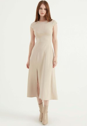 Beige aansluitende midi-jurk met korte mouwen, voorzien van een split aan de voorkant, een gladde textuur en subtiele naaddetails, gecombineerd met bijpassende enkellaarsjes.