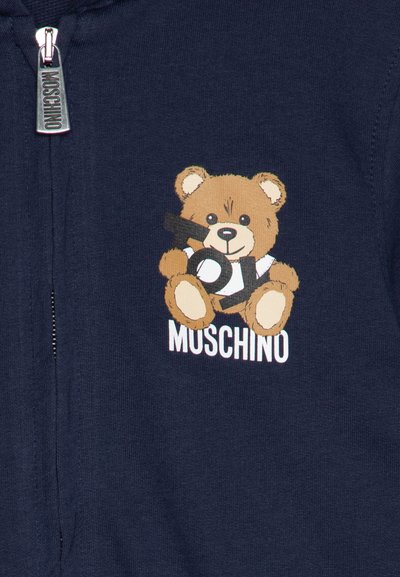 Mornarska kapuca s grafiko plišastega medvedka in detajlom besedila igrač. Zadrga, mehak material in logo "Muscino" na dnu.
