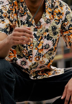 Person iført en kortærmet skjorte med orange, sort og beige blomsterprint og mørke jeans, siddende med den ene hånd hævet.