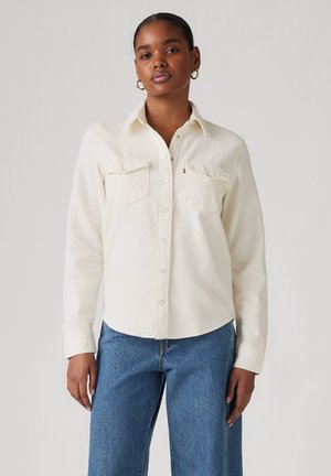 Camisa de botones de color crema hecha de suave algodón, con dos bolsillos en el pecho y herrajes plateados. Llevada con unos vaqueros de mezclilla azul y de corte holgado.