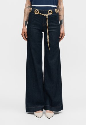 Weit geschnittene dunkelblaue Jeans aus Denim mit großen goldenen Ösen und einem goldenen Ketten-Gürtel, getragen mit weißen spitzen Schuhen und sichtbaren tätowierten Armen.