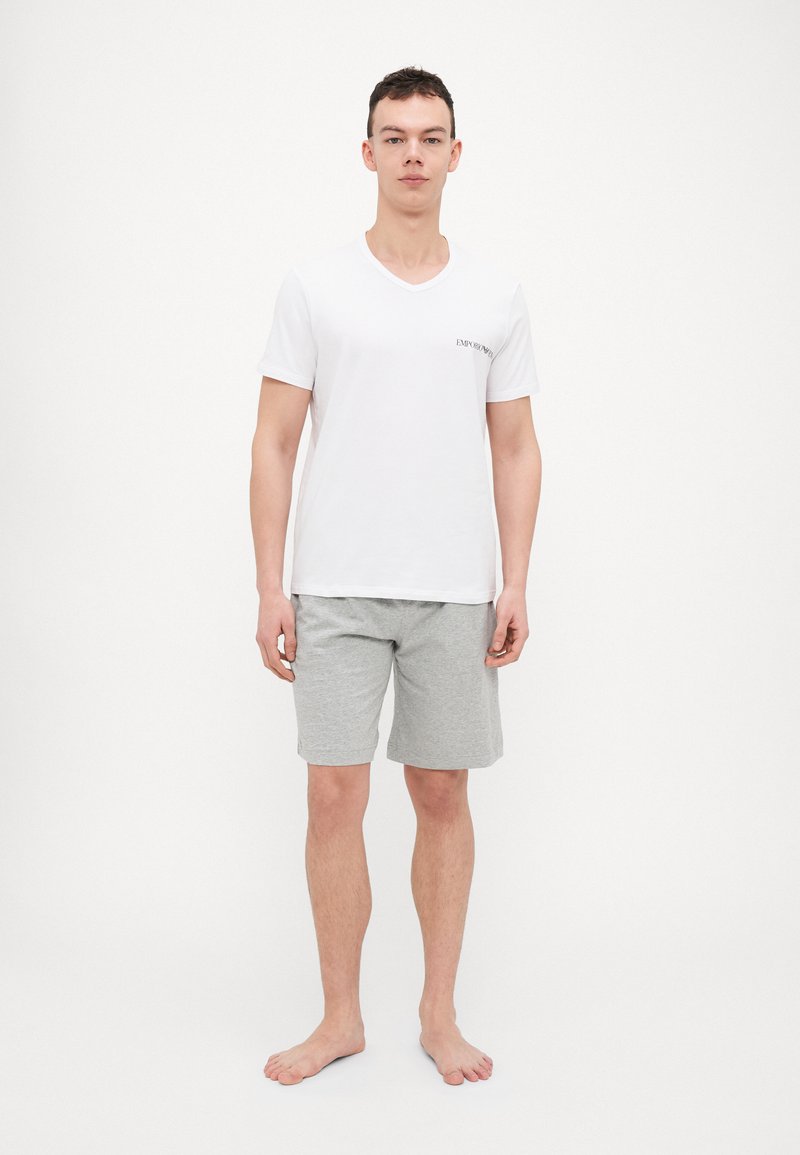 Weißes V-Ausschnitt-T-Shirt mit kleinem schwarzem Logo auf der linken Brust, kombiniert mit grauen Baumwollshorts. Fußlanger, schlichter Stoff.