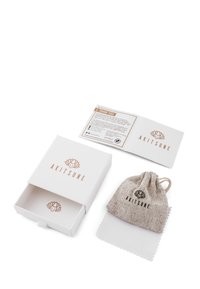 Scatola bianca con logo "AKITSUNE" in oro rosa, contenente un sacchetto in lino color beige con coulisse e un panno per la pulizia con logo. Carta delle istruzioni visibile.