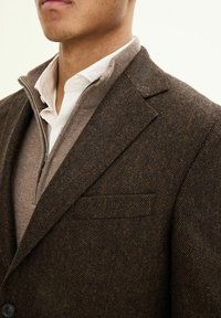 Giacca in lana con motivo a spina di pesce marrone, dotata di revers, taschino sul petto e un maglione a zip leggero color marrone chiaro sottostante.