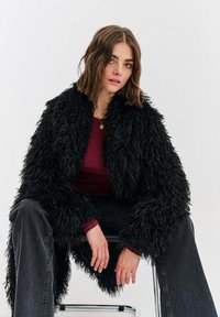 Černý kabát z umělé kožešiny s hrubou texturou, otevřený vpředu, nošený přes vínovou košili, kombinovaný s tmavými džínami.