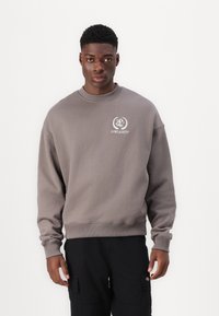 Grå genser med rund hals, ribbede mansjetter og en logo på venstre bryst. Stoffet virker mykt og teksturert.