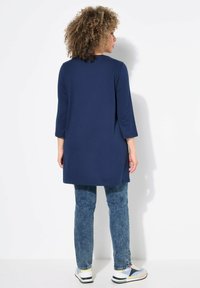 Marineblaue Tunika mit drei Viertelärmeln, gerader Schnitt und seitlichen Schlitzen, kombiniert mit hellblauen Jeans und mehrfarbigen Sneakers.