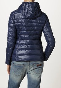 Granatowa pikowana kurtka puchowa z kapturem, z poziomymi przeszyciami, błyszczącą teksturą i dopasowanym krojem. Połączona z niebieskimi dżinsami.