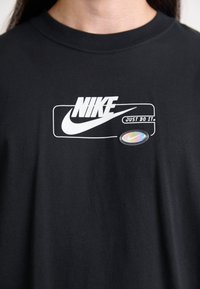 Maglietta nera con logo Nike bianco, slogan "Just Do It." e un piccolo ovale olografico con il logo swoosh Nike sul petto.