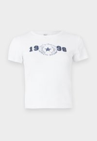 Balta kokvilnas t-krekls ar īsām piedurknēm, apaļu kakla izgriezumu, ar zilu drukāto grafiku, kurā redzams "1998" un "ATHLETIC DIVISION" ar zvaigzni.