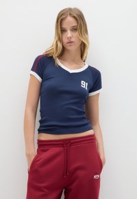 Giovane donna con capelli biondi indossa una maglietta aderente blu navy con il numero 91 e pantaloni della tuta rossi con il logo di Boston, in piedi contro un muro bianco.