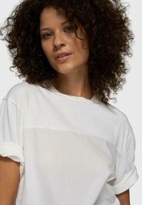 Camiseta blanca con mangas enrolladas, con una textura suave y un logo bordado sutil en el pecho. Diseño casual y de ajuste holgado.