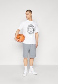 Bílá tričko s šedým grafickým motivem lva, šedé šortky, bílé ponožky a bílé tenisky, držící oranžový basketbal. Jednoduché, neformální sportovní oblečení.