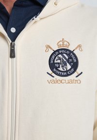 Sudadera con capucha beige cremosa con cremallera, confeccionada en tela suave y con un logo bordado en azul y dorado que incluye un caballo y coronas, con detalles en contraste en azul marino.