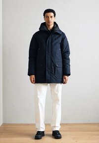 Navy blue insulated parka with a hood, two front pockets, and drawstring ties. Worn over white pants and black shoes on a wooden floor.