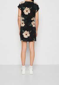 Czarna sukienka w kwiaty z krótkimi rękawami, zdobiona dużymi beżowymi kwiatami i zielonymi liśćmi. Model nosi białe trampki i skarpetki kostkowe.