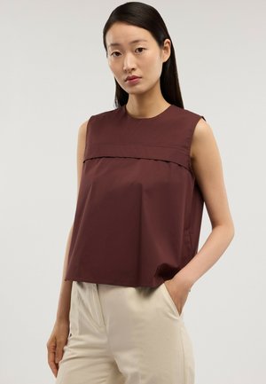 Frau mit langen dunklen Haaren, die ein ärmelloses dunkelbraunes Oberteil und hellbeige Hosen trägt, steht mit einer Hand in der Tasche vor einem schlichten Hintergrund.