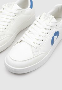 Zapatillas blancas hechas de cuero sintético suave. Cuentan con un acento azul en el talón y un logo gráfico en el lateral. Cordones planos.