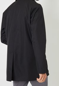 Manteau noir au design épuré, tissu imperméable, manches longues et ouverture au dos pour faciliter les mouvements. Coupe ajustée.