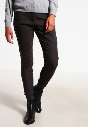 Personne portant un pantalon noir slim, des bottines noires et un pull gris, debout avec une jambe légèrement croisée sur l'autre.