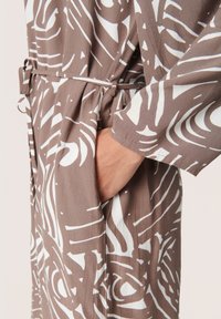 Tessuto marrone con motivo a fantasia e design astratti bianchi; presenta una vita con cintura e tasche laterali. La texture morbida e la vestibilità ampia offrono comfort.