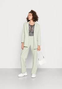 Blazer verde-claro e calças largas combinadas, feitas de tecido suave, sobre um top de padrão floral, complementados por uma mala branca e sapatos.