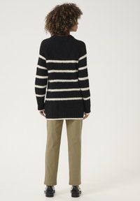 Juodai baltas dryžuotas megztinis su tekstūruotu mezgimu, apvaliu iškirpimu, ilgomis rankovėmis ir ilgesniu nugaros kraštu, derinamas su smėlio spalvos kelnėmis.