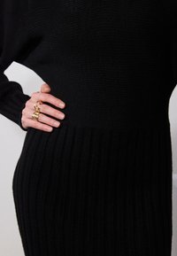 Schwarzes Strickkleid mit geripptem Design, tailliertem Schnitt und ausgestelltem Saum. Eine Hand mit einem goldenen Statement-Ring liegt auf der Taille.