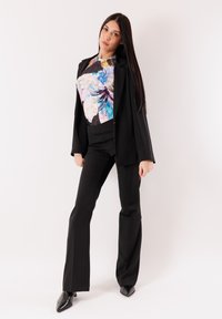 Completo nero su misura con pantaloni svasati abbinato a una maglia con motivo floreale nei toni del blu e rosa. Il modello posa su uno sfondo bianco.