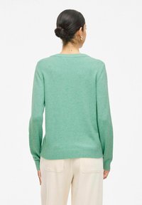 Modelo vistiendo un suéter verde y pantalones de color crema, mostrado de espaldas contra un fondo blanco.