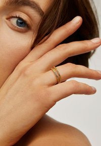 Anillos multibanda de oro con detalles texturizados, que presentan un acabado suave y pequeños acentos de color. Llevados en una mano sobre un fondo neutro.