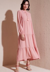 Robe maxi rose, à étages, avec des manches longues, un détail à nouer au cou et une coupe décontractée. Portée avec des sandales à talons colorées. Fond texturé.