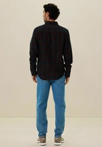 Geruite shirt in donkere kleuren met rode en blauwe accenten, met knopenkraag en lange mouwen, gedragen met lichtblauwe spijkerbroek en grijze sneakers.