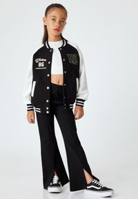 Schwarz-weiße College-Jacke mit "Whatever 86" Stickerei, kombiniert mit schwarzen Hosen mit gespaltenem Bein und schwarzen Sneakern mit weißen Sohlen.