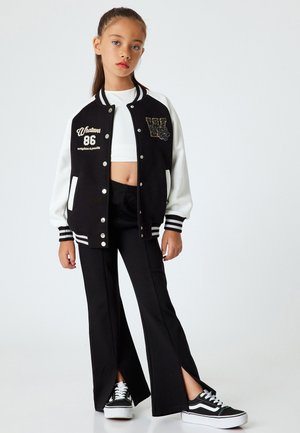Schwarz-weiße College-Jacke mit "Whatever 86" Stickerei, kombiniert mit schwarzen Hosen mit gespaltenem Bein und schwarzen Sneakern mit weißen Sohlen.