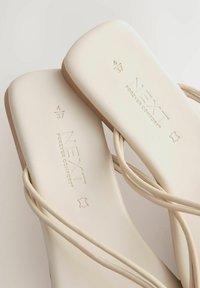 Beige sandaler med en slät, matt yta; tunna, korslagda remmar; präglat varumärke; vadderad fotsula; minimalistisk design.