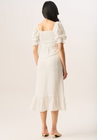White midi dress with a smocked back, puff sleeves, and a ruffled hem. Soft, textured fabric. Model wears yellow heeled sandals.
