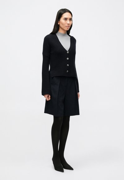 Sötétkék kötött kardigán V-nyakkivágással és öt gombbal, sötét szoknyával, fekete harisnyával és hegyes orrú fekete magassarkúval párosítva.