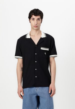Chemise noire à manches courtes avec col et poignets rayés blancs, présentant une poche poitrine avec un emblème floral. Tissu lisse avec fermeture à boutons.