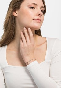 Pulsera de plata con un diseño retorcido, llevada en la muñeca. La modelo lleva una blusa blanca de manga larga con escote cuadrado.