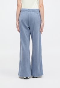 Άτομο ντυμένο με λευκά φαρδιά παντελόνια από φλις σε ανοιχτό μπλε και λευκά αθλητικά παπούτσια, standing against a plain white background, με τη πλάτη στραμμένη προς την κάμερα.