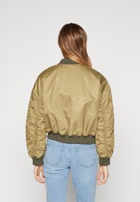 Jachetă bomber de culoare verde măslin cu un finisaj satinat, guler și tiv cu nervuri și mâneci voluminoase. Poartă cu blugi din denim de un albastru deschis.